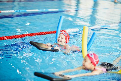Zwei Kinder mit Schwimmnudeln schwimmen im Pool, eines lächelt und hält ein Schwimmbrett in der Hand.