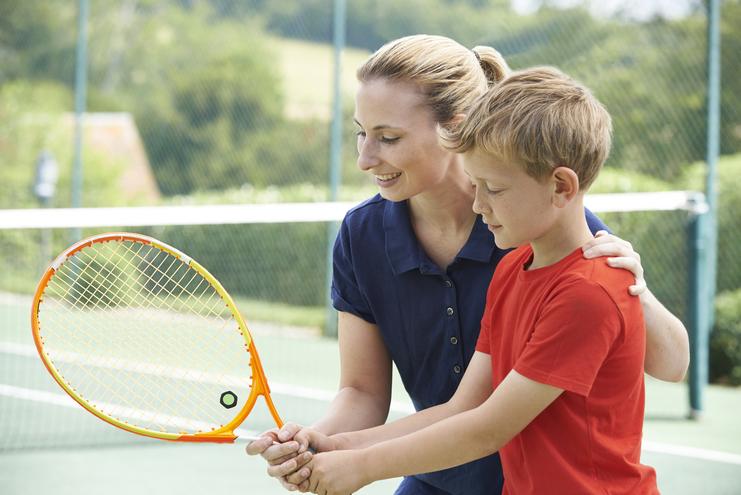 Eine Frau hilft einem Jungen beim Halten eines Tennisschlägers auf einem Tennisplatz.