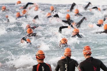 Gruppe von Schwimmern in Neoprenanzügen mit orangefarbenen Badekappen, die ins Wasser starten.